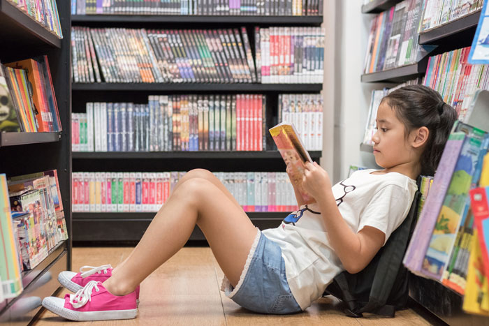 A girl is sitting on the floor leaning against a bookshelf and surrounded by books as she reads a book.